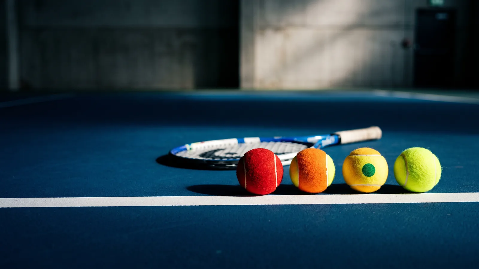 Red, orange, green-dot and yellow tennis balls in a row on a blue court beside a junior racquet